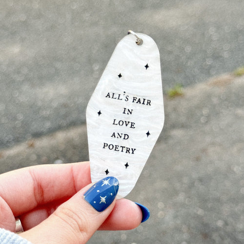 A white marble acrylic keychain with the quote "All's fair in love and poetry" inspired by Taylor Swift written in black. Black sparkles are scattered around the motel keychain.