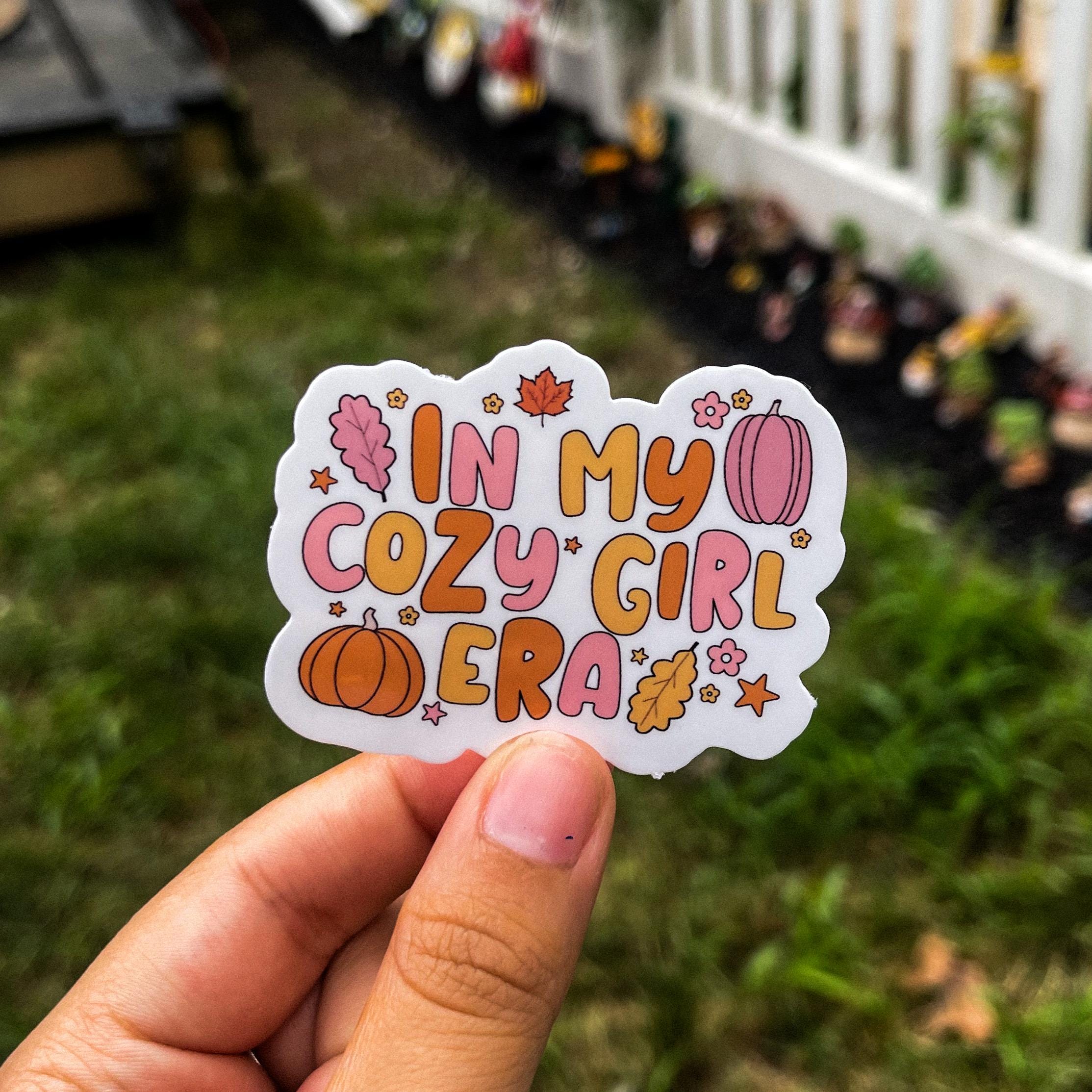 Aesthetic fall sticker with the quote "in my cozy girl era" surrounded by leaves, pumpkins, flowers, and stars. the overall color scheme is orange, pink, and yellow, alternating between each object.