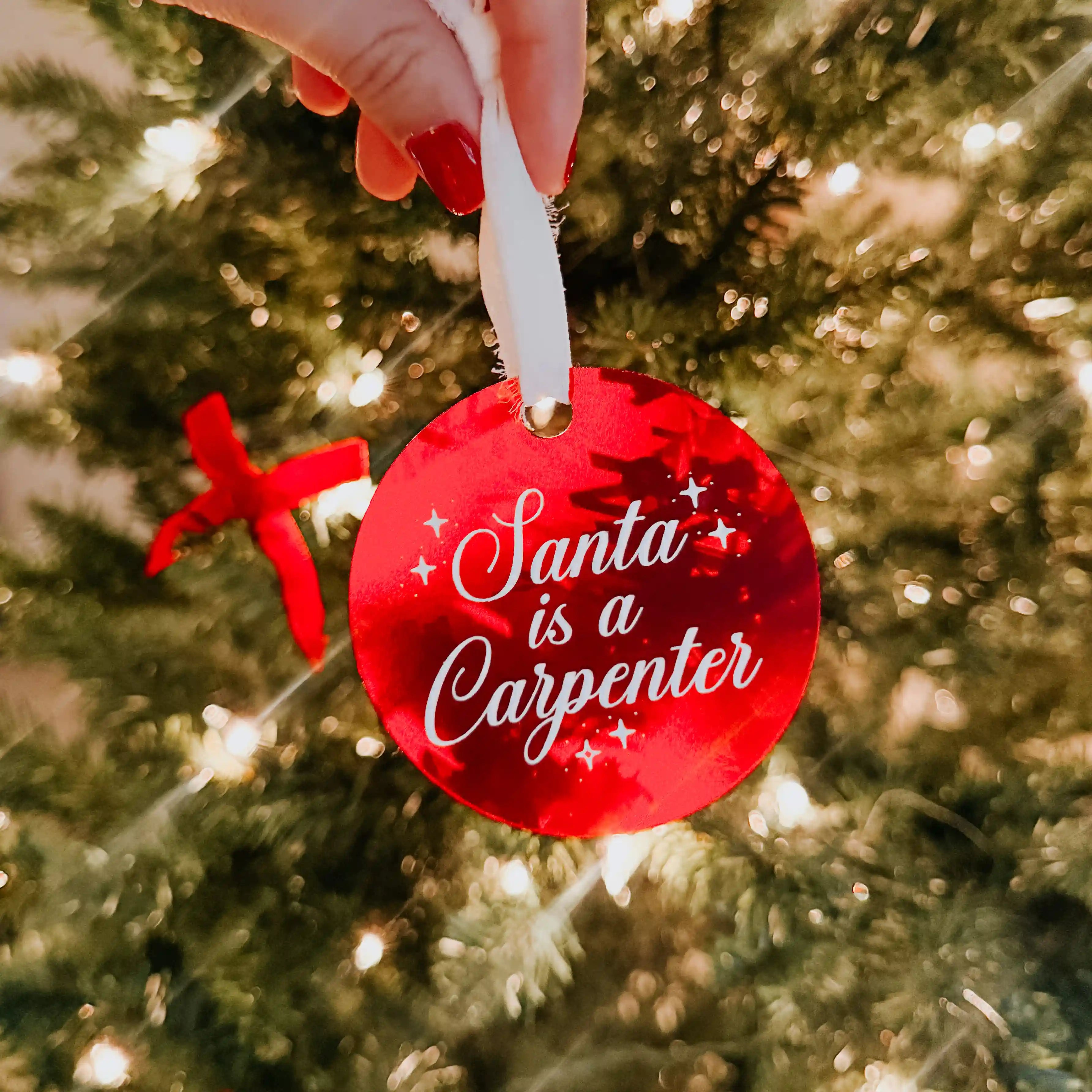 A red Sabrina Carpenter inspired Christmas ornament with the quote "Santa was a Carpenter" engraved and painted on the front and tied with a white chiffon ribbon. 