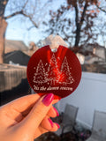 Round circle ornament made with frosted red acrylic, with a white chiffon ribbon tied at the top. Engraved and painted white on the front of the ornament are three Christmas trees, little sparkles, and the lyrics "'tis the damn season"