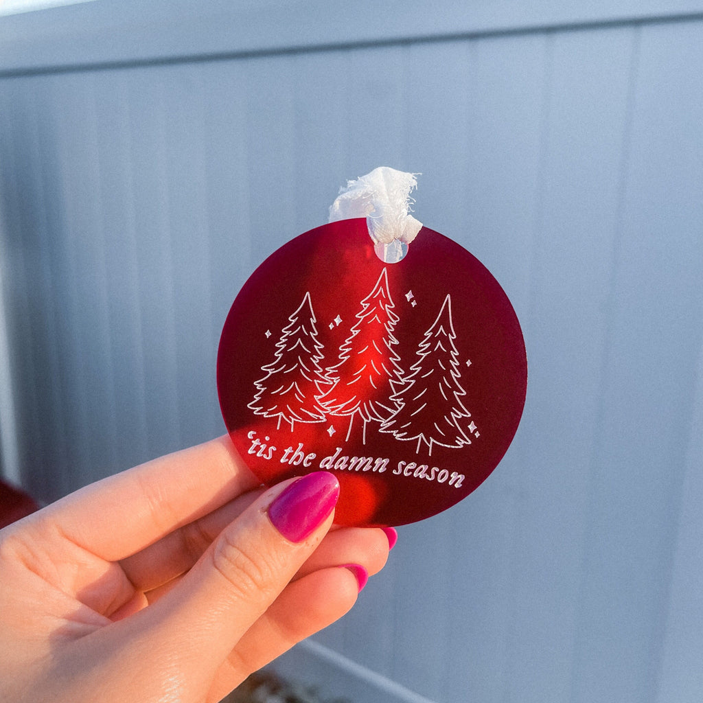 Round circle ornament made with frosted red acrylic, with a white chiffon ribbon tied at the top. Engraved and painted white on the front of the ornament are three Christmas trees, little sparkles, and the lyrics "'tis the damn season"