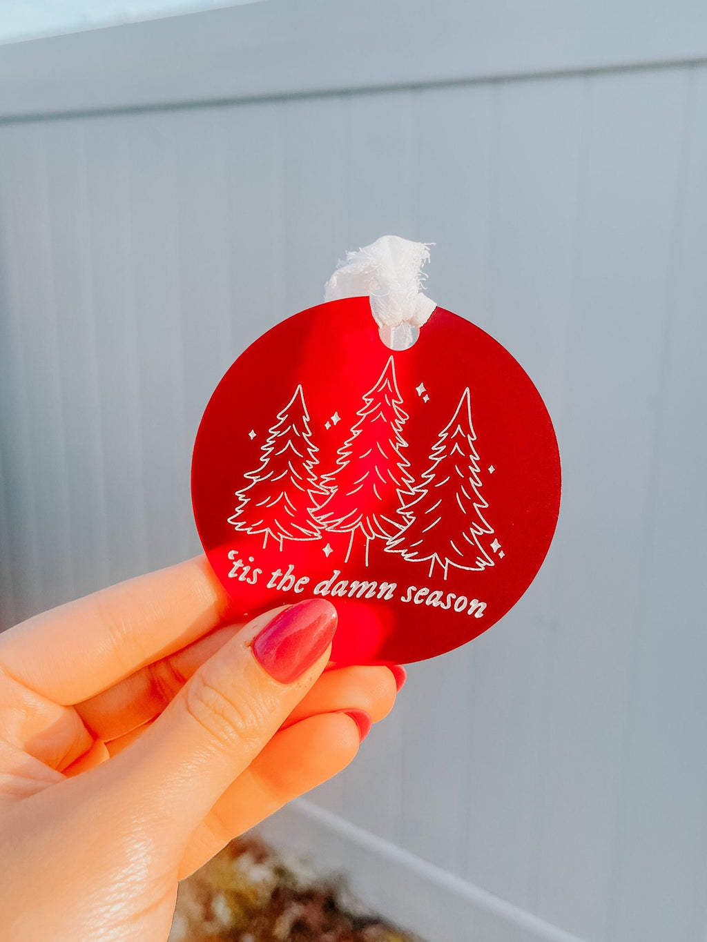 Round circle ornament made with frosted red acrylic, with a white chiffon ribbon tied at the top. Engraved and painted white on the front of the ornament are three Christmas trees, little sparkles, and the lyrics "'tis the damn season"