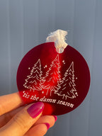 Round circle ornament made with frosted red acrylic, with a white chiffon ribbon tied at the top. Engraved and painted white on the front of the ornament are three Christmas trees, little sparkles, and the lyrics "'tis the damn season"
