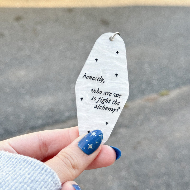 White marble acrylic motel keychain engraved with black sparkles and the text "Honestly, who are we to fight the alchemy".
