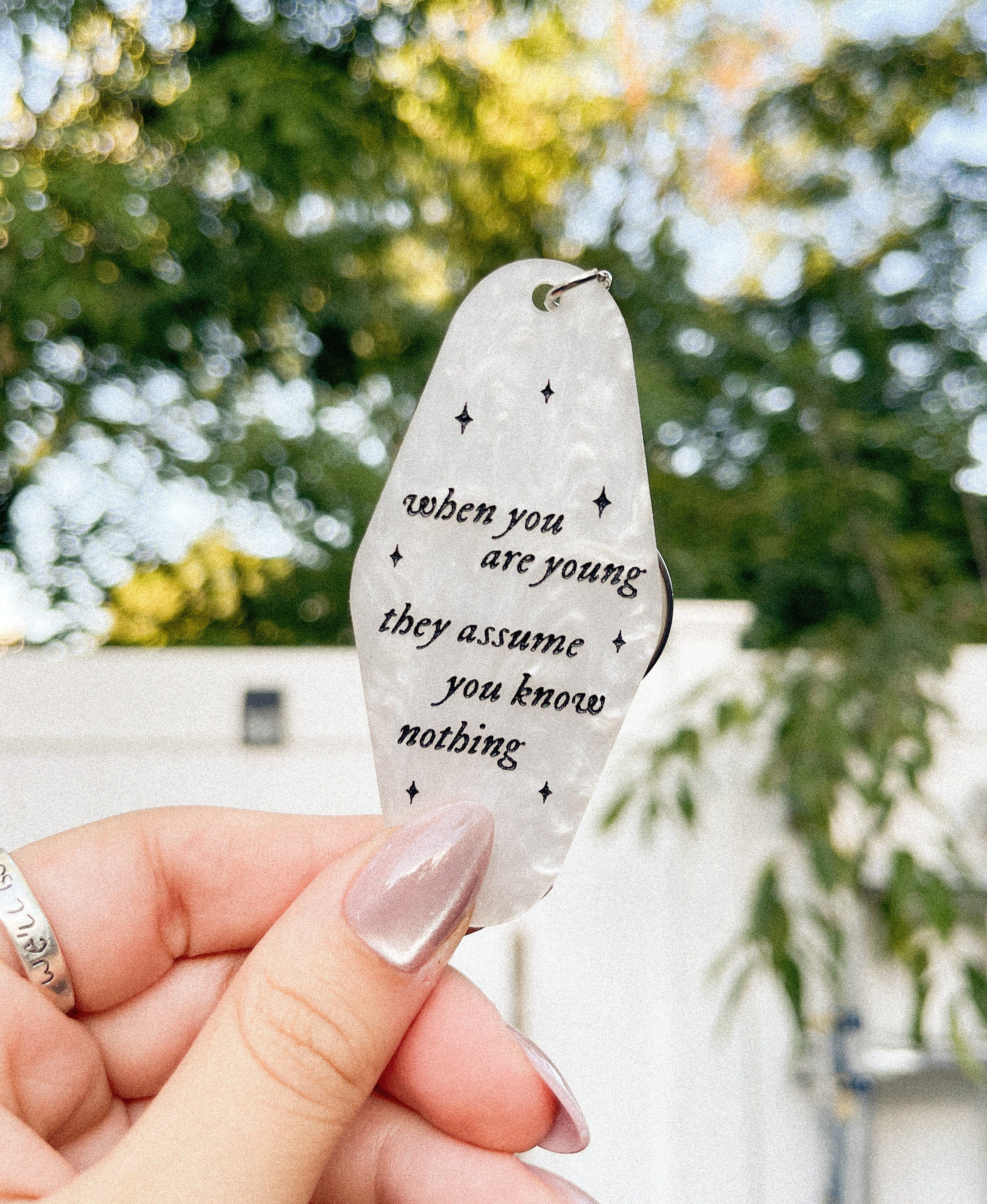 A hand holding a white marble acrylic motel keychain engraved with black sparkles and the text "when you are young they assume you know nothing"