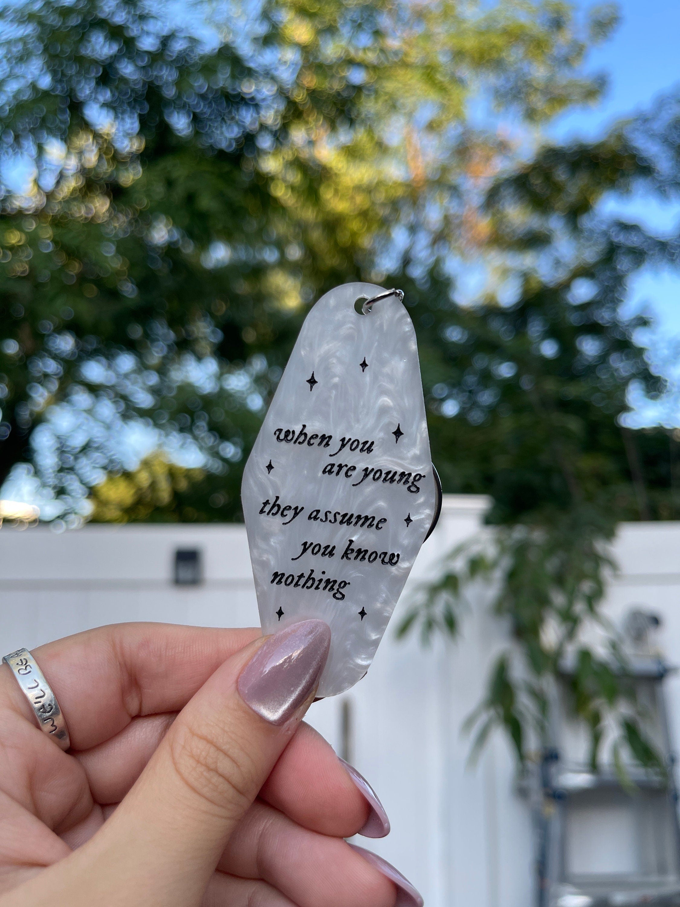 A hand holding a white marble acrylic motel keychain engraved with black sparkles and the text "when you are young they assume you know nothing"