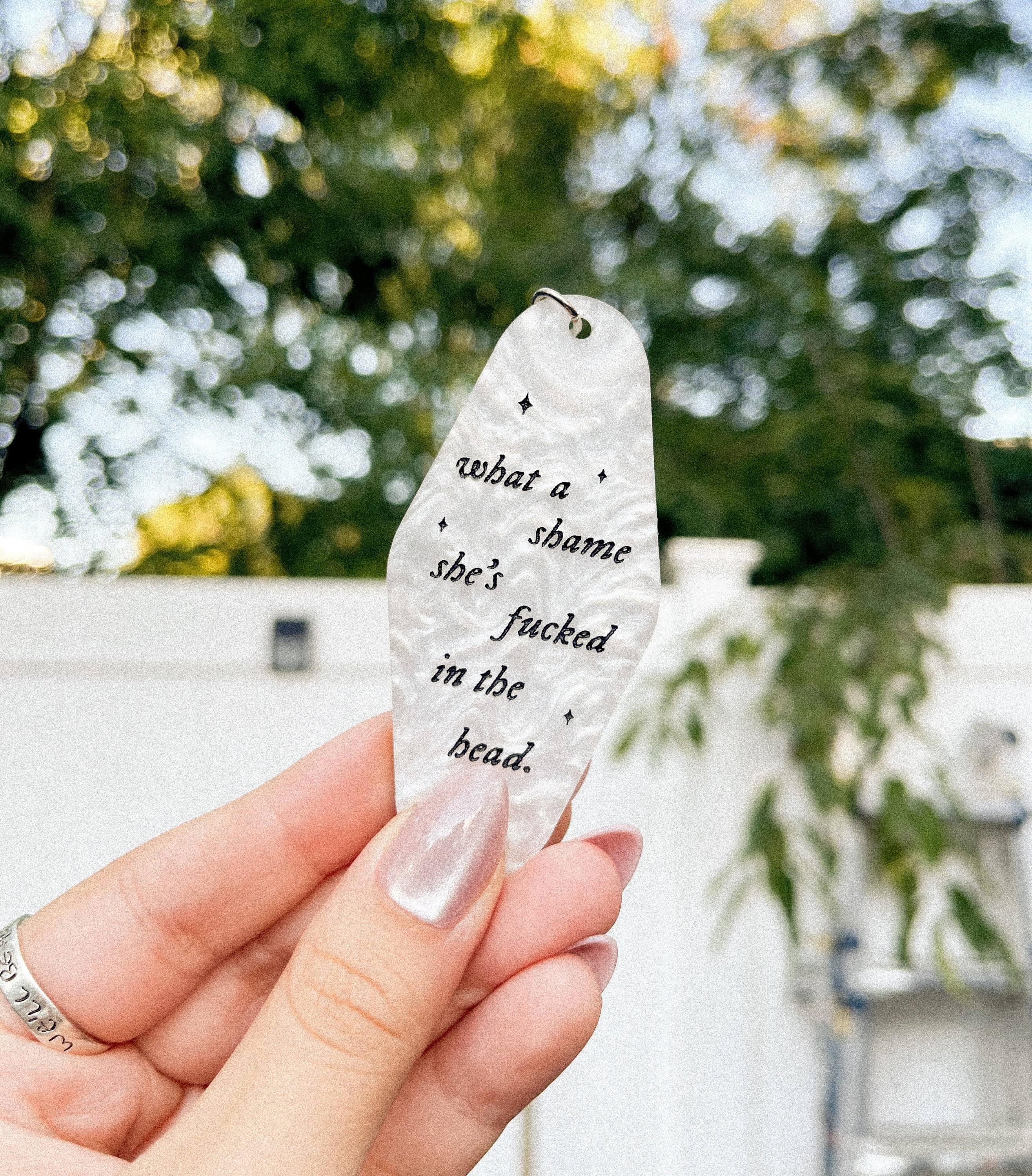 A hand holding a white marble acrylic motel keychain engraved with black sparkles and the text "what a shame she's fucked in the head"