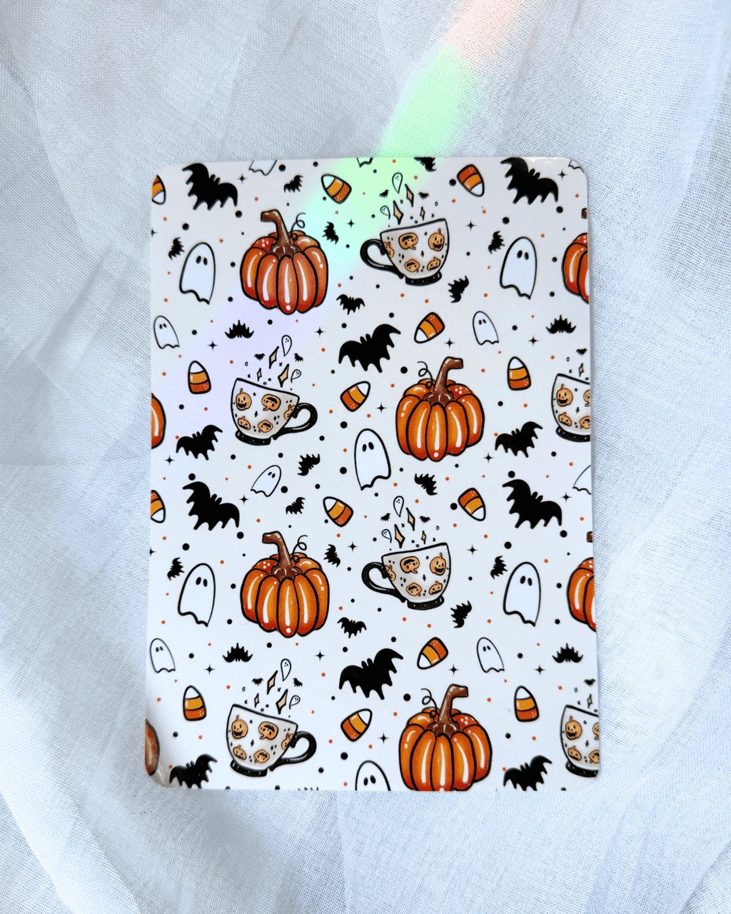 A kindle insert with a Halloween-themed pattern featuring orange pumpkins, black bats, white ghosts, and orange-and-white coffee cups, displayed on a white tulle fabric background with decorative fairy lights.