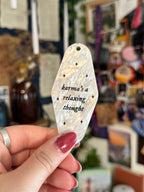 White marble acrylic motel keychain engraved with black sparkles and the lyrics "karma's a relaxing thought.'