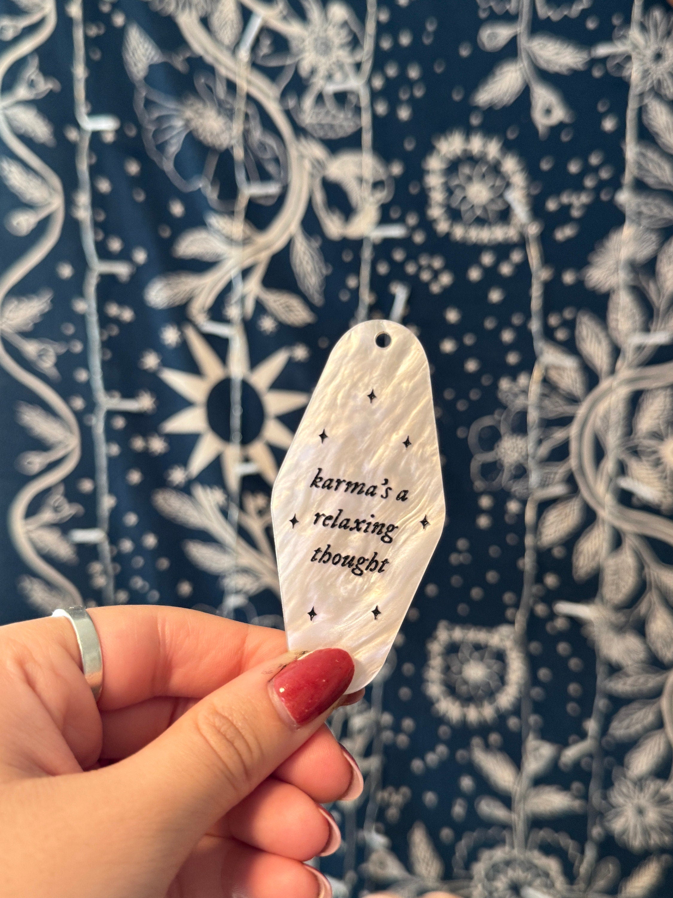White marble acrylic motel keychain engraved with black sparkles and the lyrics "karma's a relaxing thought.'