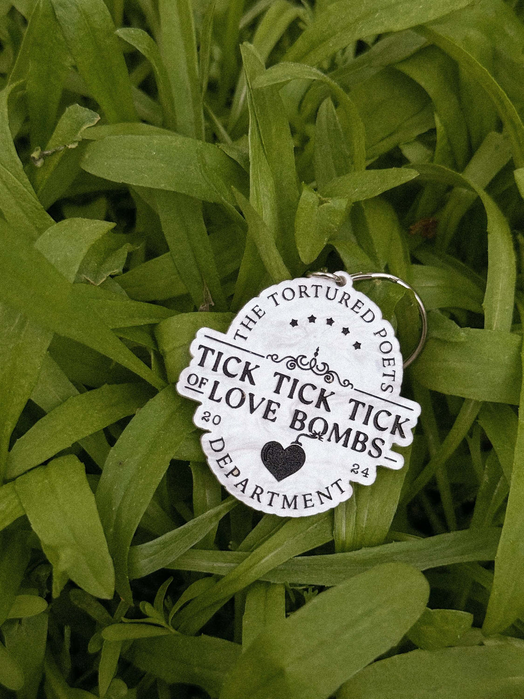 A white marble die-cut acrylic keychain with black text reading 'TICK TICK TICK OF LOVE BOMBS' and 'THE TORTURED POETS DEPARTMENT' photographed lying on green grass.