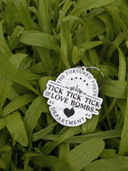 A white marble die-cut acrylic keychain with black text reading 'TICK TICK TICK OF LOVE BOMBS' and 'THE TORTURED POETS DEPARTMENT' photographed lying on green grass.