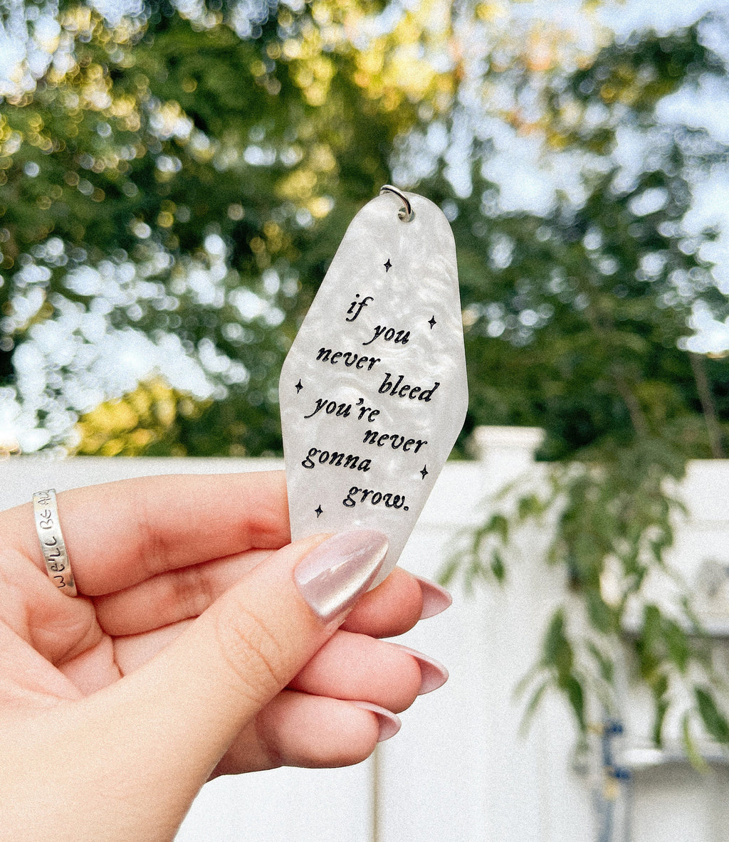 A hand holding a white marble acrylic motel keychain engraved with black sparkles and the text "if you never bleed you're never gonna grow"