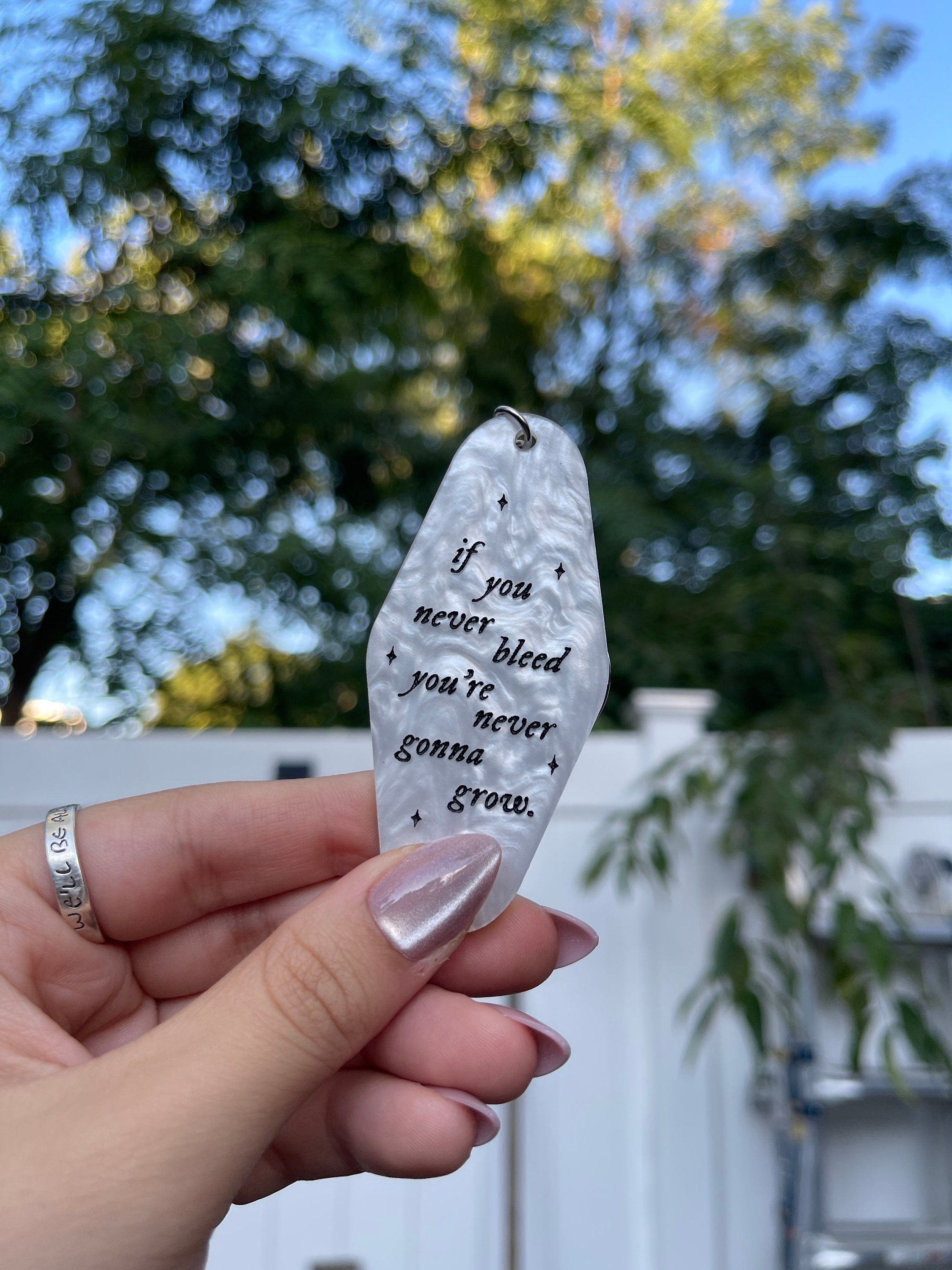 A hand holding a white marble acrylic motel keychain engraved with black sparkles and the text "if you never bleed you're never gonna grow"