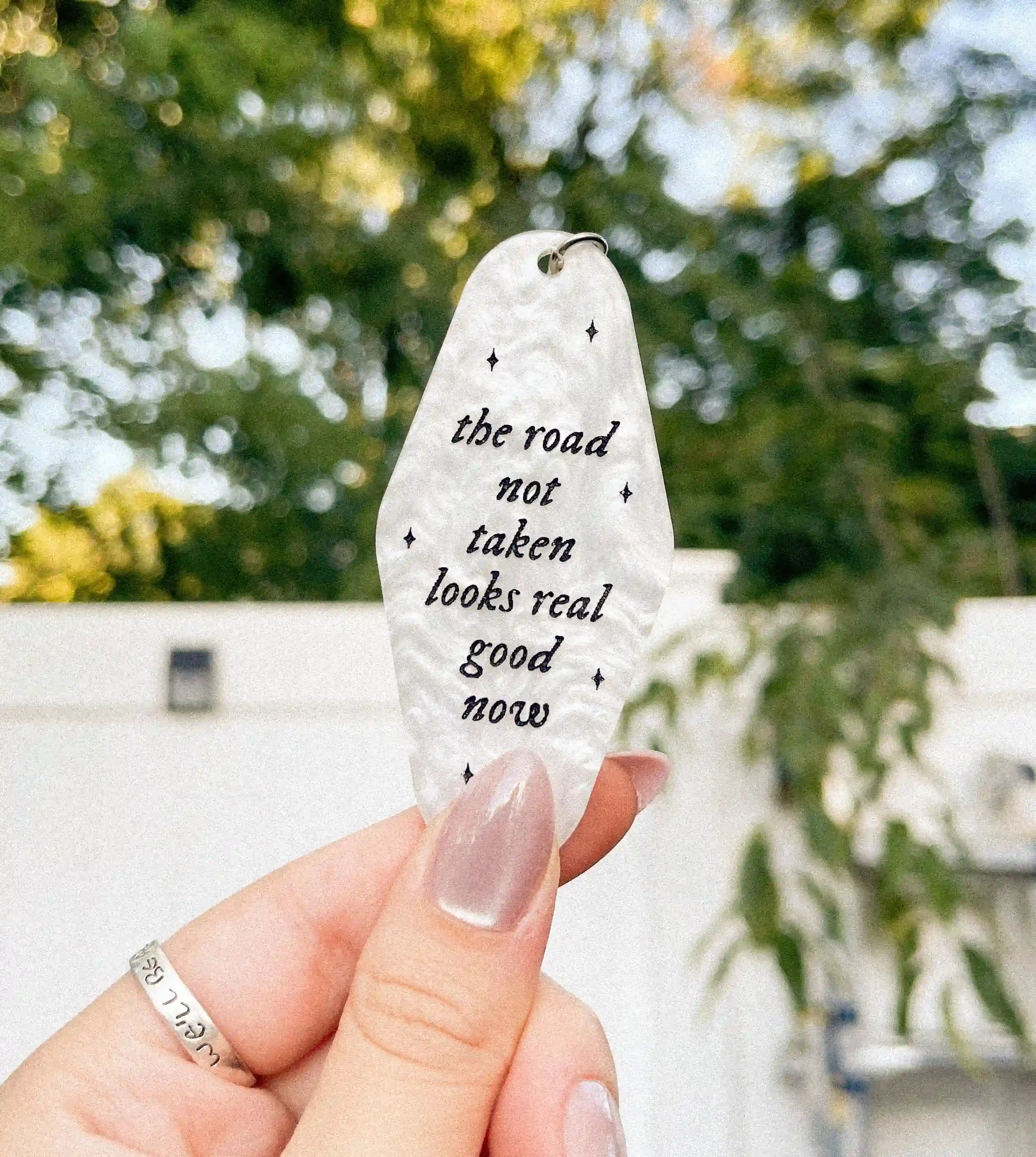 A hand holding a white marble-colored keychain with the text 'THE ROAD NOT TAKEN LOOKS REAL GOOD NOW' engraved on it inspired by taylor swift.
