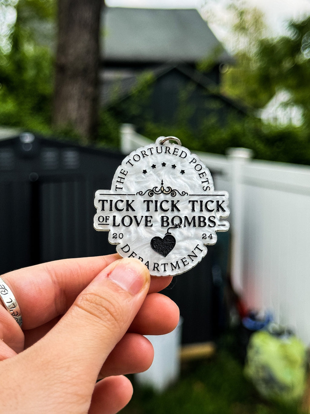 A hand holding a white marble acrylic keychain with black engraved text "The Tortured Poets Department 2024. Tick tick tick of love bombs."